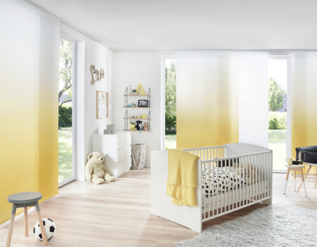 Helles Kinderzimmer mit Flächenvorhang in weiß bzw. mit Farbverlauf zu gelb und Blick in den Garten, Kinderbett und Kommode, Teppich, Stuhl, Teddybär und Regal mit Dekoration